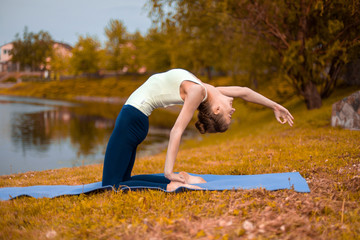 Fototapeta premium Slim brunette girl goes in for sports and performs yoga poses in the fall in nature by the lake