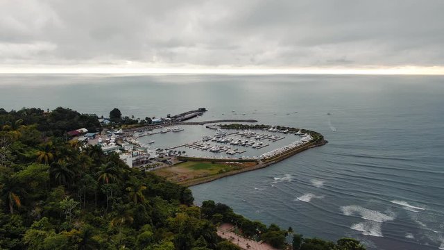 Drone Footage of Marina Pez Vela near Quepos, Costa Rica on a Cloudy Day with Blue Open Ocean in the Background, Camera Dolly-In Pushing Passed Green Tropical Jungle Hill