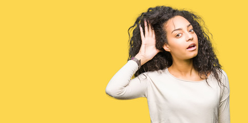 Young beautiful girl with curly hair wearing casual sweater smiling with hand over ear listening an hearing to rumor or gossip. Deafness concept.