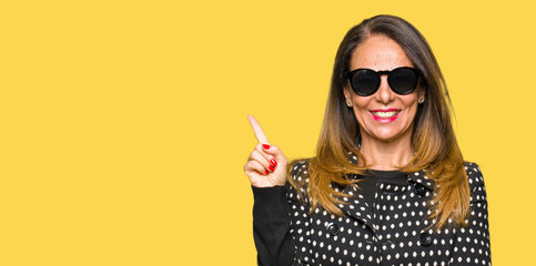 Beautiful middle age woman wearing sunglasses with a big smile on face, pointing with hand and finger to the side looking at the camera.