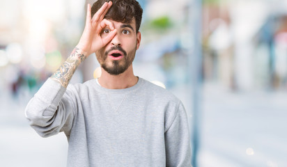 Young handsome man wearing sweatshirt over isolated background doing ok gesture shocked with surprised face, eye looking through fingers. Unbelieving expression.