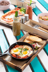 Substantial israeli breakfast on summer terrace: shakshuka with tomatoes, salami, chili pepper and fried eggs, served in hot cast pan with toasts