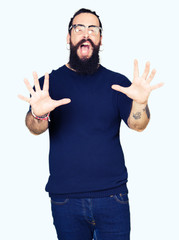 Young hipster man with long hair and beard wearing glasses afraid and terrified with fear expression stop gesture with hands, shouting in shock. Panic concept.