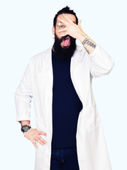 Doctor therapist man with long hair and bear wearing white coat peeking in shock covering face and eyes with hand, looking through fingers with embarrassed expression.