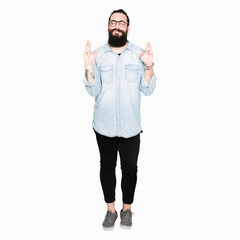 Young hipster man with long hair and beard wearing glasses smiling crossing fingers with hope and eyes closed. Luck and superstitious concept.
