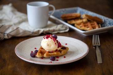 Close up of delicious crunchy waffle with ice cream and berries with coffee and more waffles in the background, wooden table setting