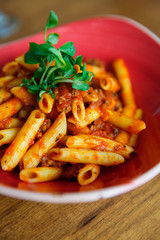 close up of a plate of penne bolognese with a small pile of green bean sprouts on top on a wooden table with a glass of water in the background by the window of a restuarant