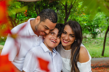 couple of Latin American men and women, with boy suffers autism, happy in a portrait family outdoors together in a park, the three laughing hugging