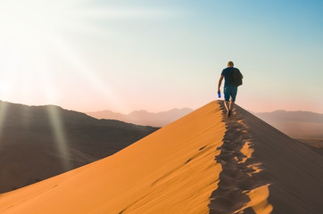 A man with backpack goes up on sand dune in sunset. Desert landscape