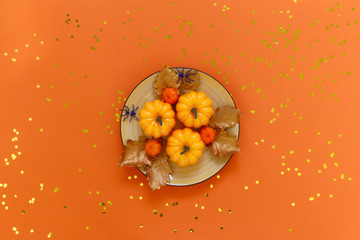 Pumpkins on a yellow dish with spiders  on orange background with golden confetti. Flat lay, top view fall autumn and halloween concept.