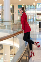 young white girl in red jacket, heels and black trousers leans on railing in mall and looks away. Full height.