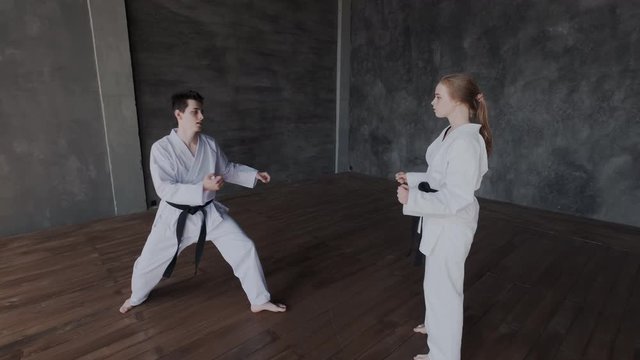 A dark-haired guy and a blonde girl are training martial arts in the gym. Sparring or single duels in taekwondo school. Jump kick attack. Fight stance. Overall, general view.