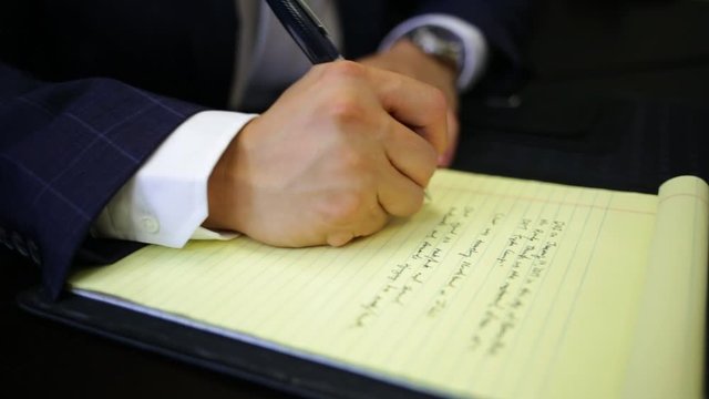 Hands of man in suit taking notes on legal pad.