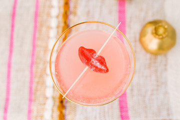 Pink fruity sparkling champagne cocktail with bubbles served in gold rimmed coupe glass, garnished with sugary candy lips on a bohemian woven striped fabric surface / valentine and romance concept