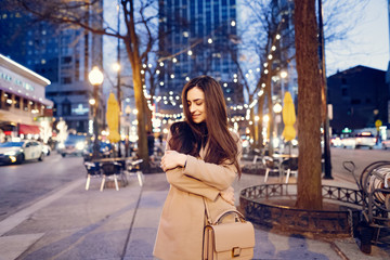 Fototapeta premium Beautiful girl in a evening city. Stylish brunette in a brown coat. Woman in a Chicago. Lady with a phone