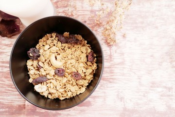 Granola, a kind of breakfast cereal consisting typically of rolled oats, brown sugar or honey, dried fruit, and nuts, in a black bowl on wood background. There is a glass of milk is nearby.