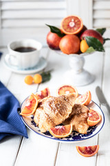 Breakfast croissants lie on a blue plate and red oranges on a white background. Orange juice. A cup of coffee