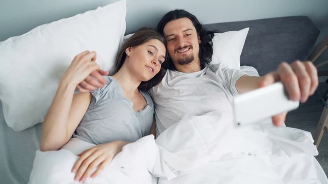 Bearded guy is taking selfie in bed with beautiful girlfriend using smartphone camera having fun together. Modern technology, photo and bedroom concept.