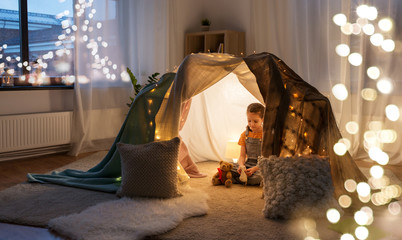 childhood and hygge concept - happy little girl with toys in kids tent at home