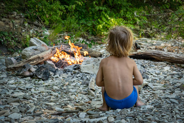 cute little boy in the forest sitting near the fire in the summer, view from the back