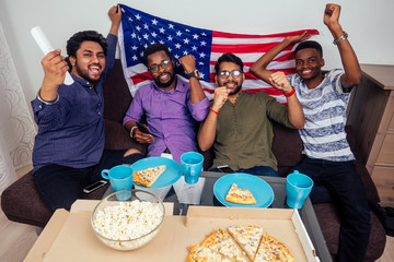 african american males eating pizza and popcorn , cheering and smiling while watching TV match at home