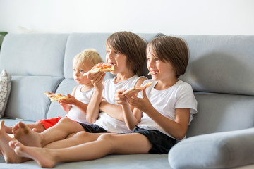 Obraz na płótnie Canvas Cute children, sitting on couch, eating pizza and watching TV. Hungry child taking a bite from pizza on a pizza party