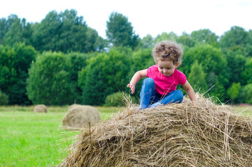 Naklejka na ściany i meble Cute girl sitting on hay stack or bale on yellow wheat field in on sunny summer day. Baby play at hay bales during summer harvest time in Germany during Oktoberfest. soft focus.