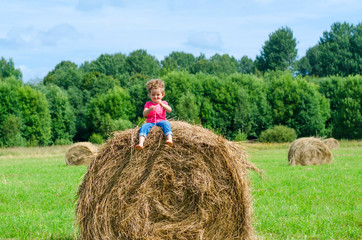 Naklejka na ściany i meble Little girl sitting on hay stack or bale on yellow wheat field in on sunny summer day. Baby play at hay bales during summer harvest time in Germany during Oktoberfest. soft focus.