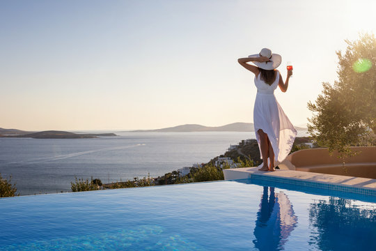 Frau mit wei&szlig;em Hut steht am Pool und genie&szlig;t die Aussicht auf das Meer bei Sonnenuntergang mit einem Aperitif in der Hand