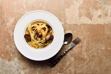 top view of delicious pasta with mollusks served with spoon and knife on weathered beige background