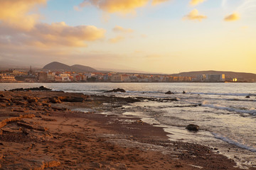 Fototapeta na wymiar Golden overcast sunrise over El Medano town, in Tenerife with the limestone coast in the foreground. Mellow light sunrise over the Atlantic with calm waters, rocky shores and the town in the distance.