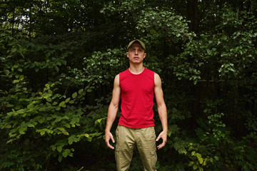 A young man in a red t-shirt, cap and pants on a background of green foliage in the forest.