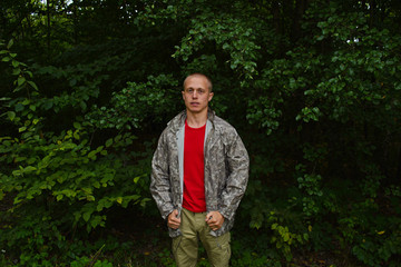 A young man in a camouflage raincoat in the forest against the green foliage.