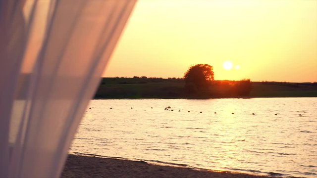 Tilt in a beach tent water front at the sunset. Sheer fabric and sea landscape  at golden hour with colorful sky. Sun Lens flare effect.