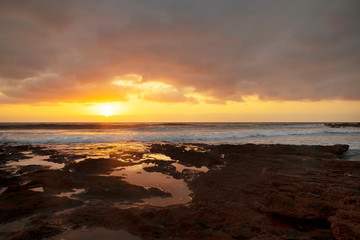 Glorious sunrise reflected in the natural water pools formed in the limestone shores of El Medano resort, Tenerife, Canary Islands, Spain, vibrant seascape with overcast sky and golden haze light