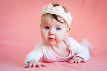 Cute healthy blue eyed Caucasian infant baby girl with a textile crown on her head on a pink background. Isolated close up image, adorable child looking at the camera.