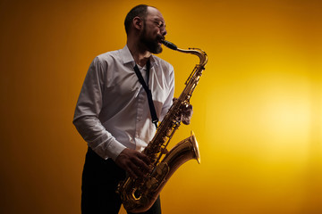 Fototapeta na wymiar Portrait of professional musician saxophonist man in white shirt plays jazz music on saxophone, yellow background in a photo studio, side view