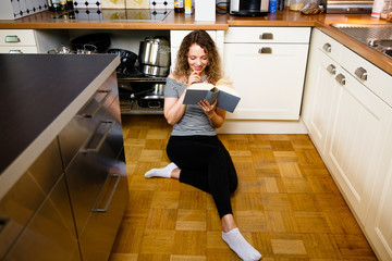 Fototapeta na wymiar Hübsche junge Frau liest ein Buch in der Küche, sitzend auf dem Fußboden.