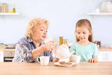 Obraz na płótnie Canvas Cute little girl and grandmother drinking tea with cookies in kitchen
