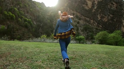 Beautiful young woman traveler runs freely across the field in nature among the mountains. Throws a hat into the air. Enjoys life, freedom and travel. Wearing a plaid shirt and hat.
