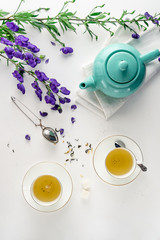 Two white tea cups with green tea, teal colored tea pot, purple flowers on white background