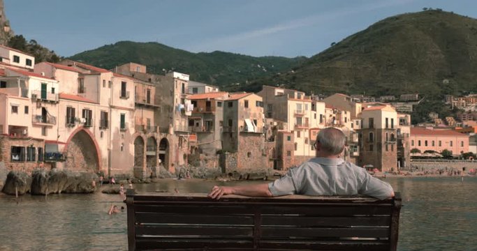 Old man is looking a seaside city in Italy sitting on a bench, sicilia island 
