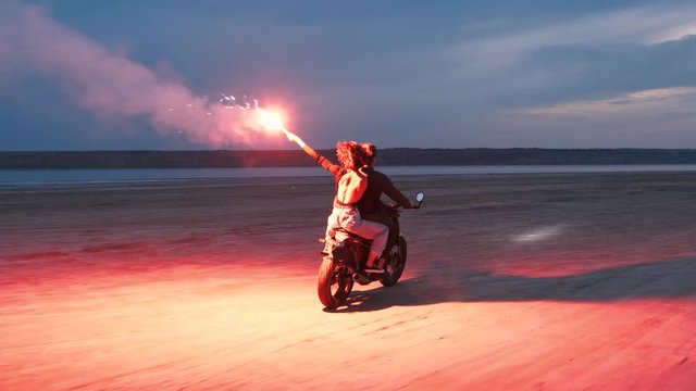 Couple riding on vintage motorcycle with red burning signal fire after sunset on beach, slow motion