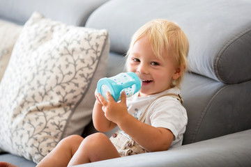 Adorable baby boy drinking milk from a bottle in a white sunny living room.
