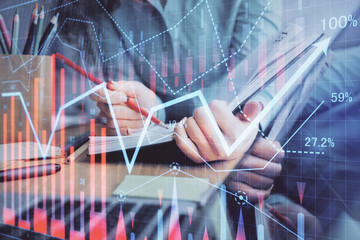 A woman hands writing information about stock market in notepad. Forex chart holograms in front....