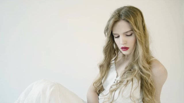 Girl with long hair in white long dress sits on white background in studio. Delicate facial features, nude makeup. Wind blows hair. She touches her hair, straightens her hair. 