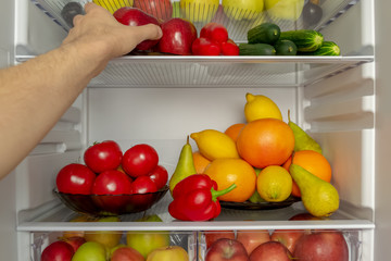 Fridge is filled with useful products. Vegetables and fruits. A man's hand takes a red Apple from the refrigerator. Diet, vegetarianism, weight loss concept