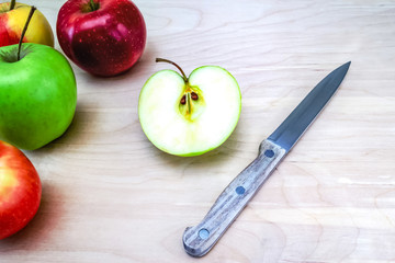 Fresh, red, green and yellow red apples, in the center is a cut apple and a knife with a wooden handle, the background is a table made of light oak