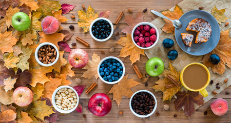 Seasonal autumn background. Frame of maple leaves and a cake, berries, raisins, apples, fruits, coffee and nuts over wooden background.