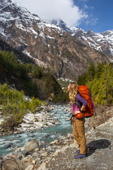 Beautiful tourist girl with backpack looking at scenic view of the valley of the Himalayas with mountain river and snow-capped mountains on Annapurna Circuit Trek in Nepal